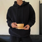 Person wearing a black hoodie holding a beige cup and saucer.
