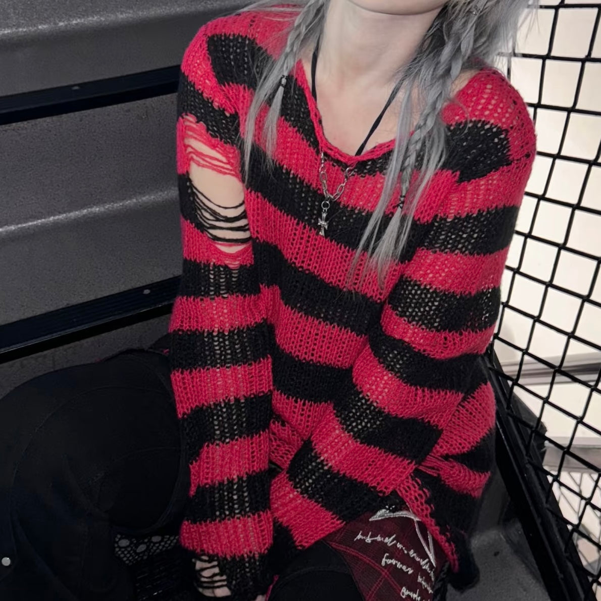 A person wearing a red and black striped distressed knit sweater, with gray hair and layered necklaces, sitting in a dark environment next to a black wire mesh screen.