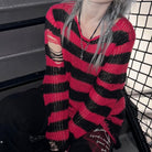 A person wearing a red and black striped distressed knit sweater, with gray hair and layered necklaces, sitting in a dark environment next to a black wire mesh screen.