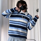 A femboy wearing a blue, navy, and white horizontally striped knit sweater over a white collared shirt, paired with gray bottoms, standing in front of white curtains.