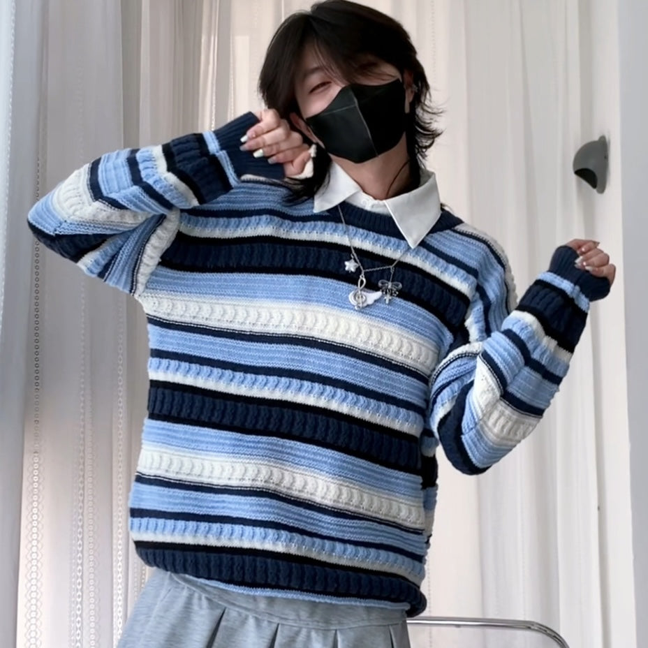 A femboy wearing a blue, navy, and white horizontally striped knit sweater over a white collared shirt, paired with gray bottoms, standing in front of white curtains.