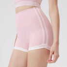 A close-up of a model wearing high-waisted light pink athletic shorts with white trim and side stripes, paired with a matching pink sports bra against a light gray studio background.