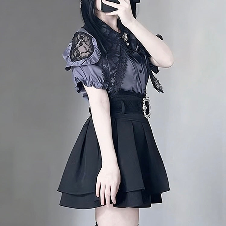 A femboy wearing a black and gray gothic Lolita-style outfit, featuring a short puff-sleeve top with lace shoulder details and a pleated black skirt, photographed against a neutral gray background.