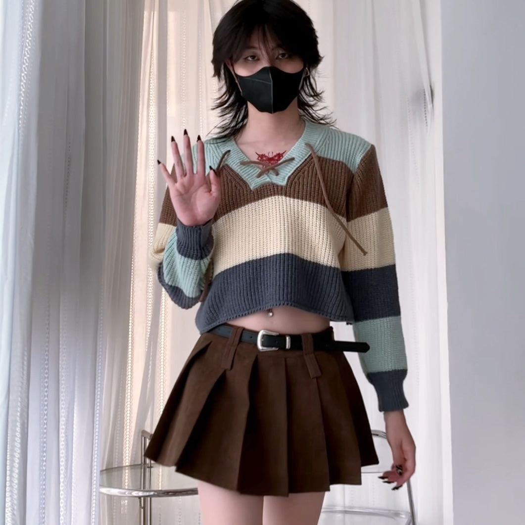 A femboy wearing a v neck lace up color block sweater with light blue jeans, seated on a dark couch.