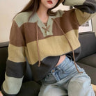 A femboy wearing a v neck lace up color block sweater with light blue jeans, seated on a dark couch.