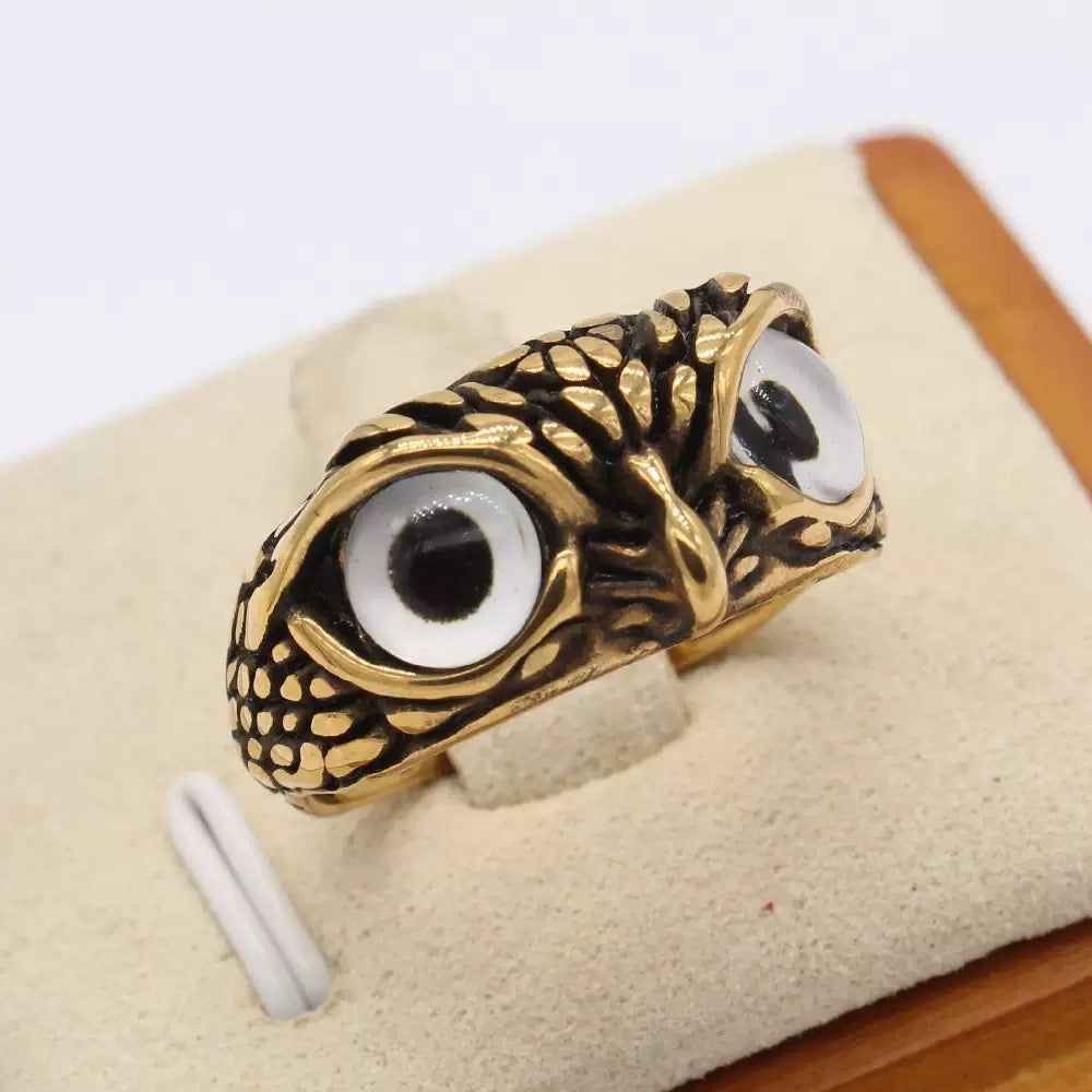 Gold and black eye-shaped ring on a beige cushion with a white background