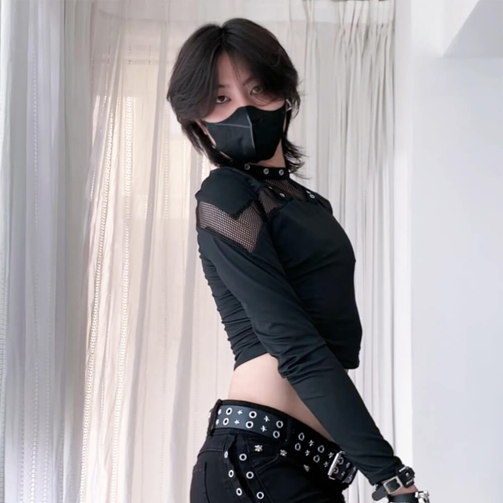 Person wearing a black outfit with sheer sleeves and studded details against a white curtain background