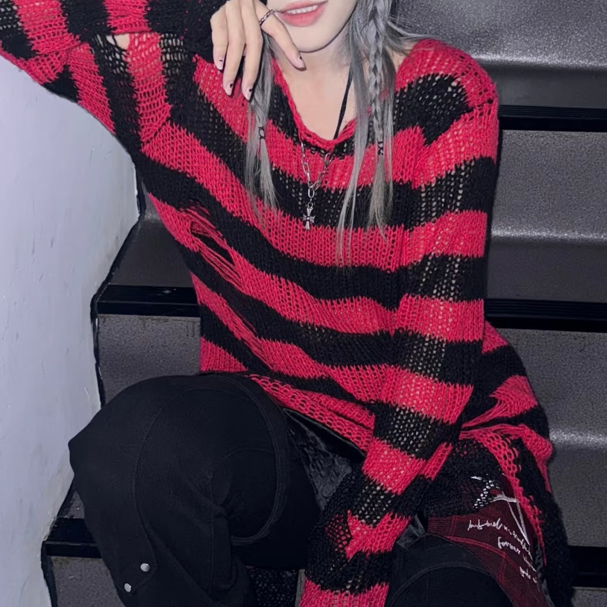 A person wearing an oversized red and black striped knit sweater, paired with black pants, sitting on dark steps.