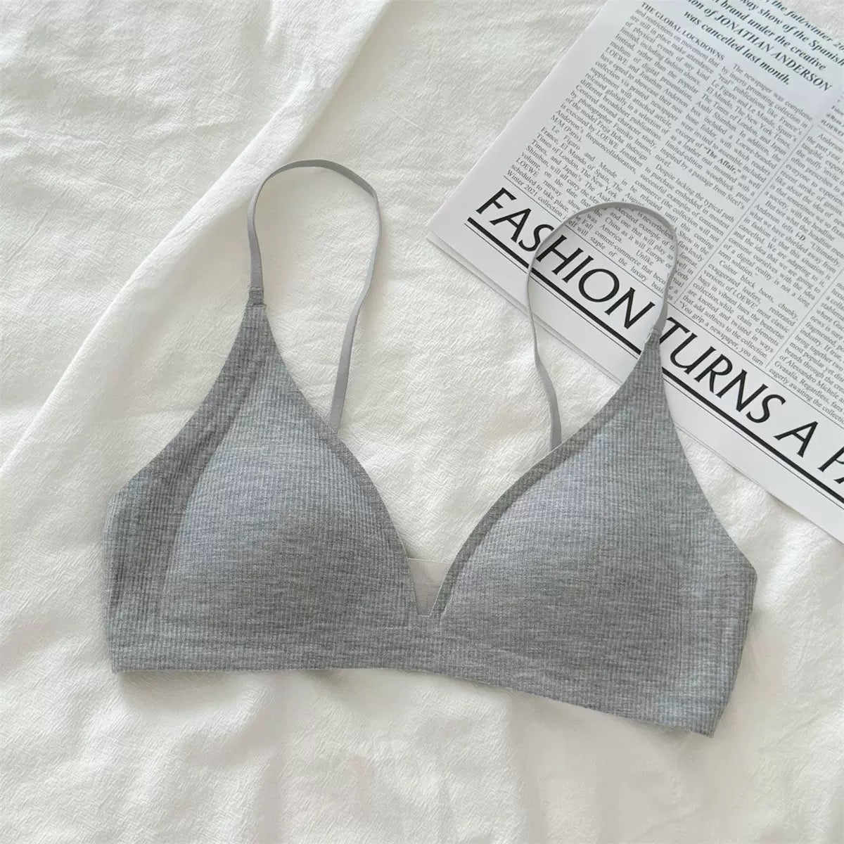 A light gray ribbed triangle bralette with thin straps, laid flat on a white textured fabric surface next to a printed magazine page.