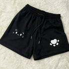 Black athletic shorts with a white cat face graphic on one leg and a white paw print graphic on the other, laid flat on a white faux fur background.