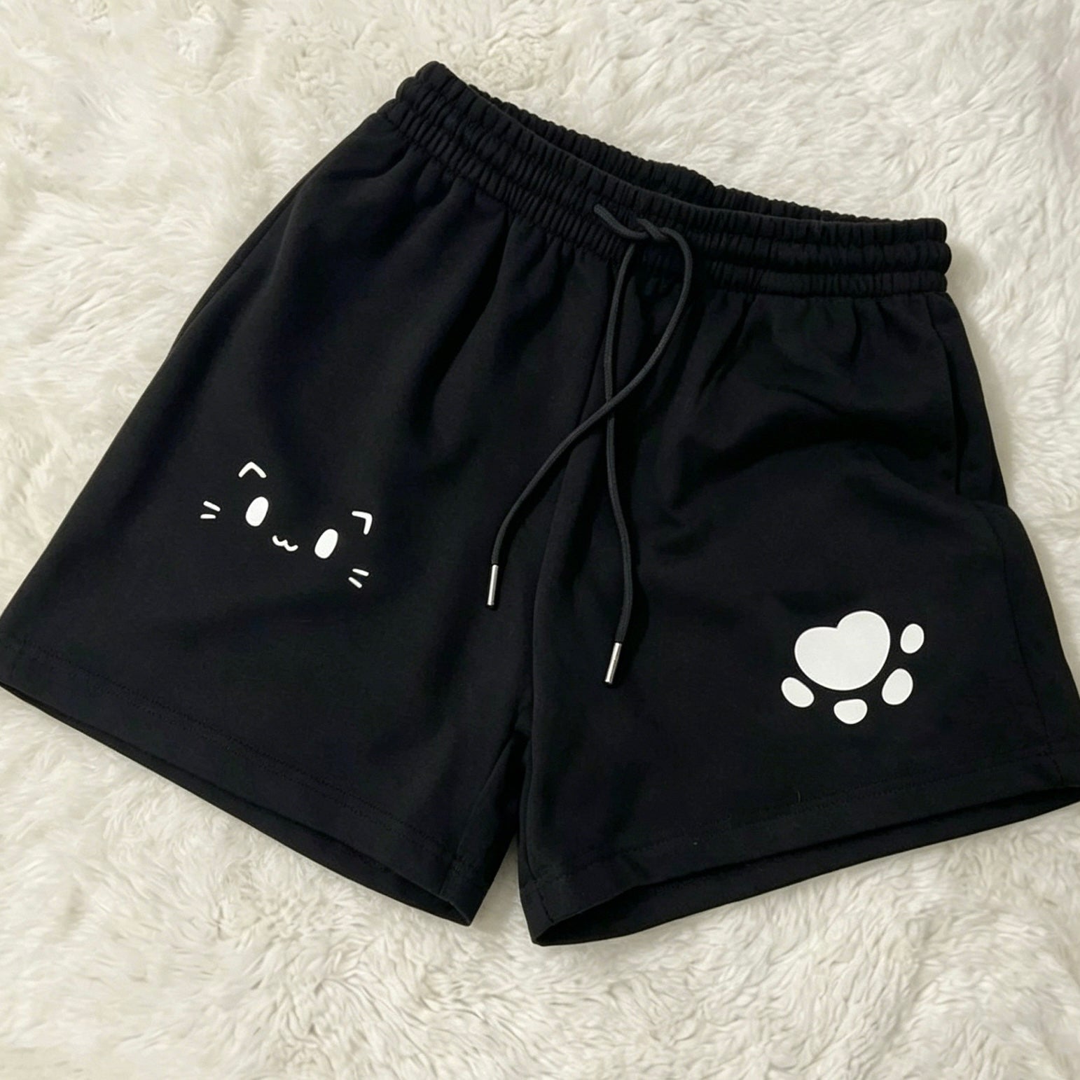Black athletic shorts with a white cat face graphic on one leg and a white paw print graphic on the other, laid flat on a white faux fur background.