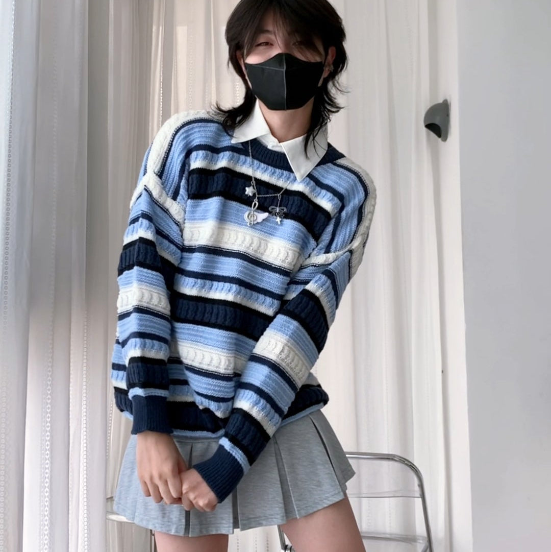 A femboy wearing a blue, white, and navy striped long-sleeve knit sweater with a white collar, paired with a light gray pleated mini skirt, white socks, and a black face mask, standing in a room with white curtains and a metal chair.