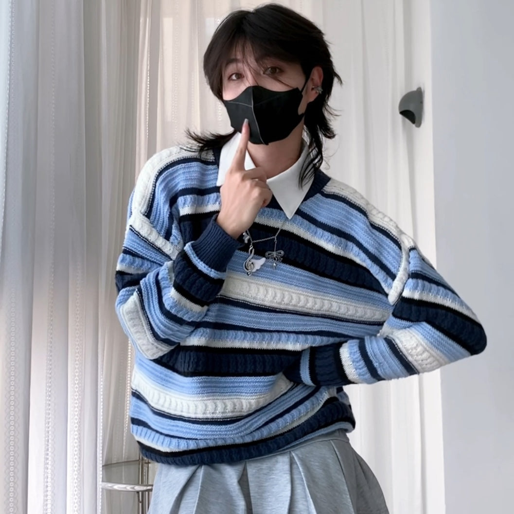 A femboy wearing a blue, navy, and white striped knit sweater over a white collared shirt, with a black face mask and gray pleated skirt, standing in front of white curtains.