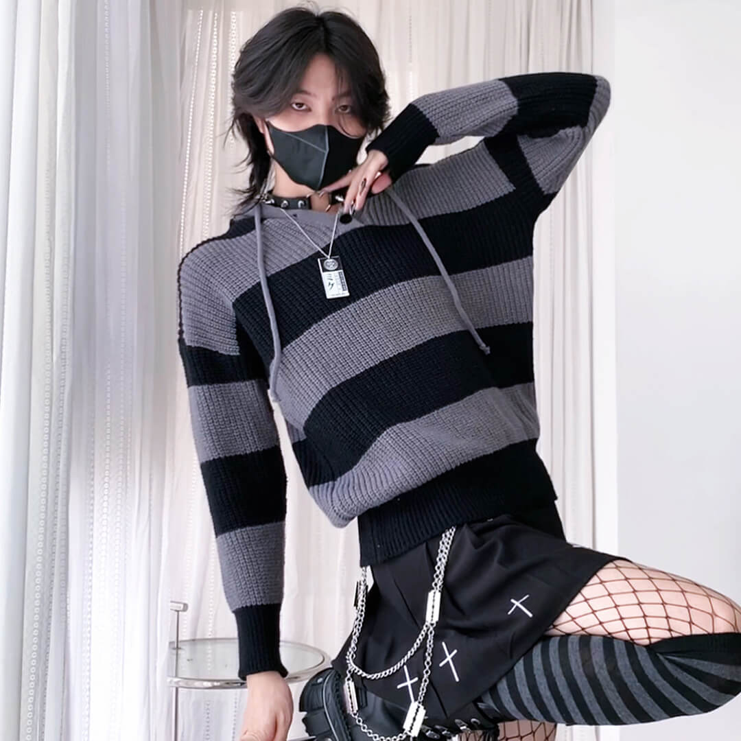 Person wearing a black and gray striped sweater with a hood, standing against a white curtain background.