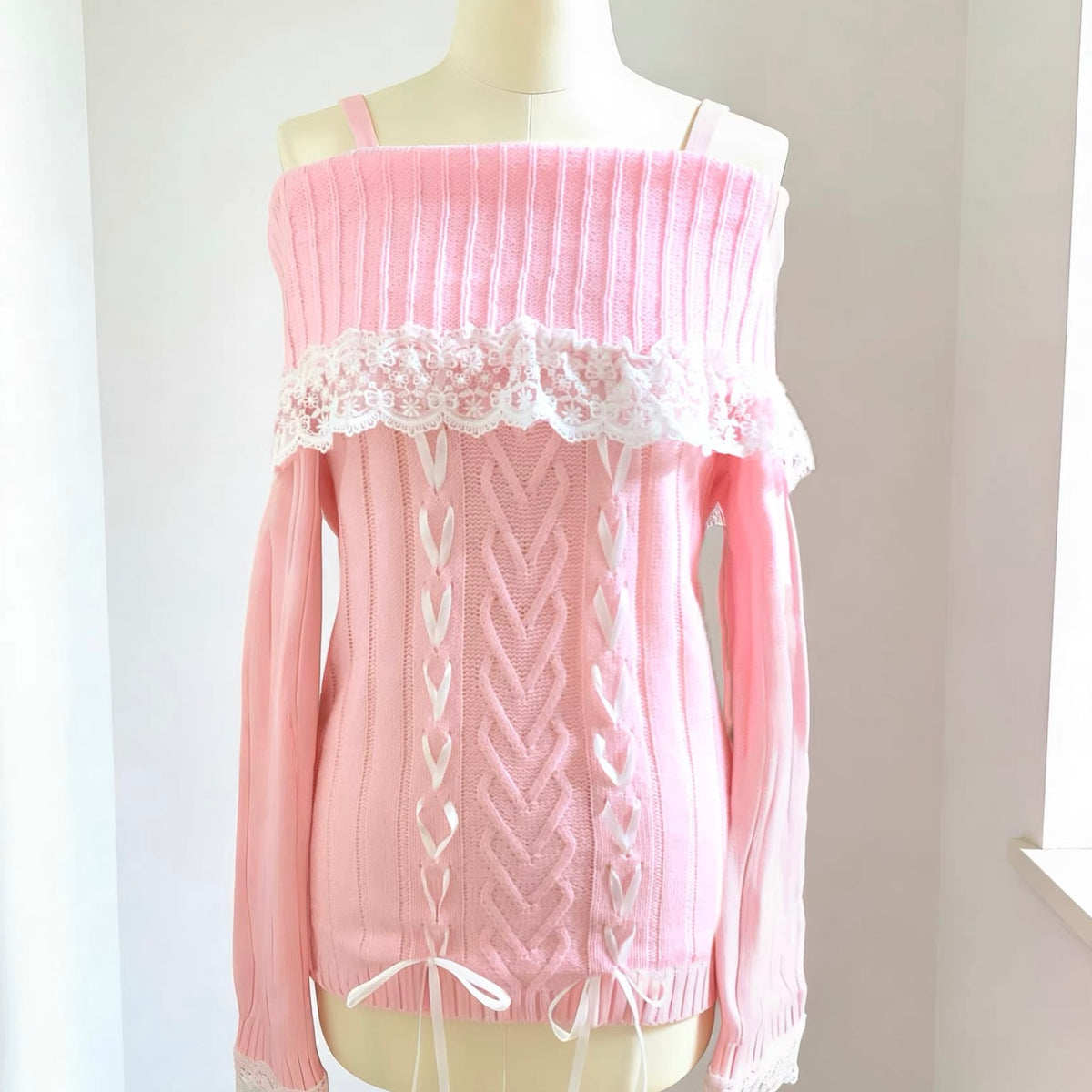 A pink off-the-shoulder cable-knit sweater with white lace trim and white lace-up front details, displayed on a cream mannequin torso against a light neutral background.