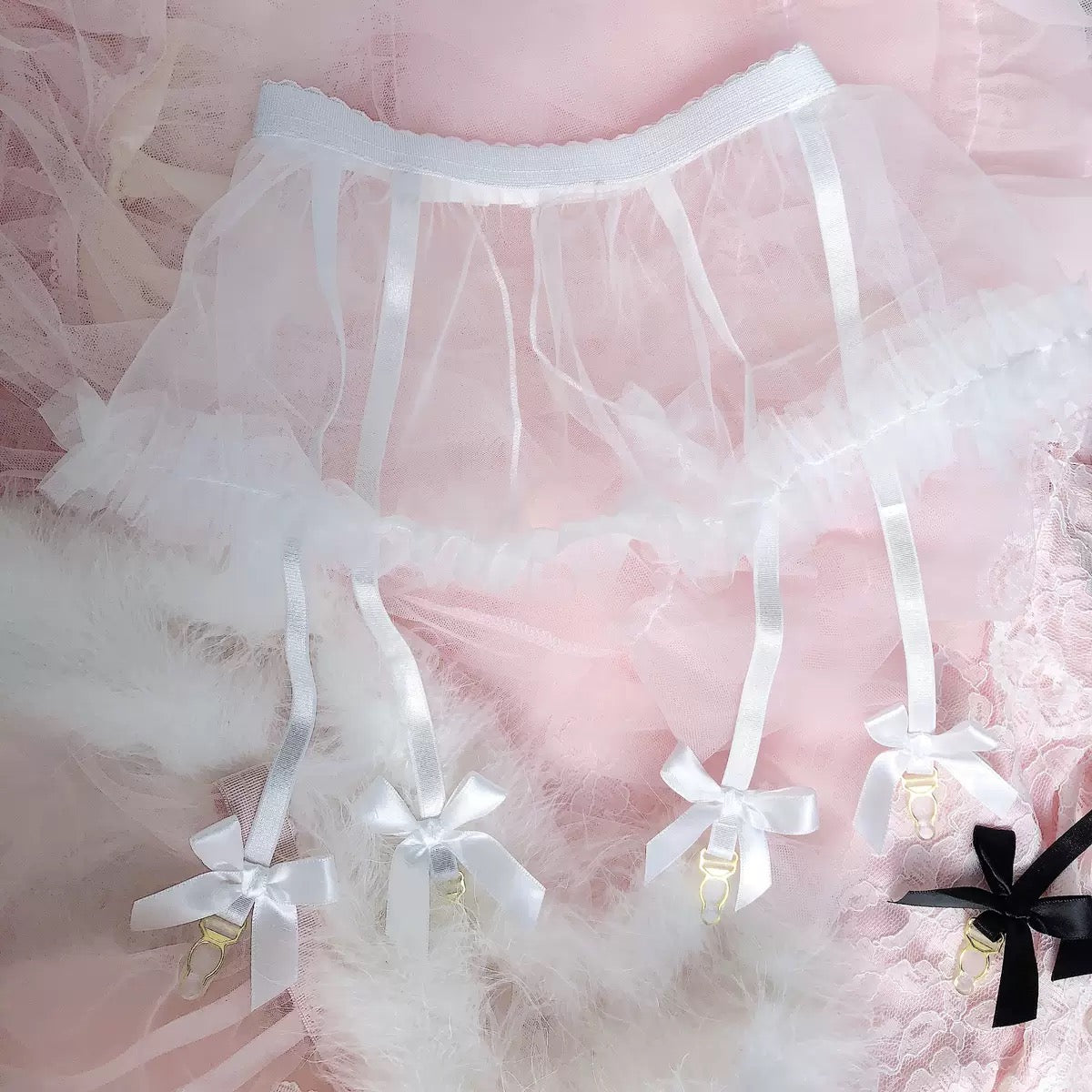White tulle garter belt with satin ribbons, bow details, and gold-tone clips, displayed on a pink lace and feather background.