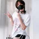 Femboy wearing a white t-shirt with cartoon design and black shorts, making peace signs.