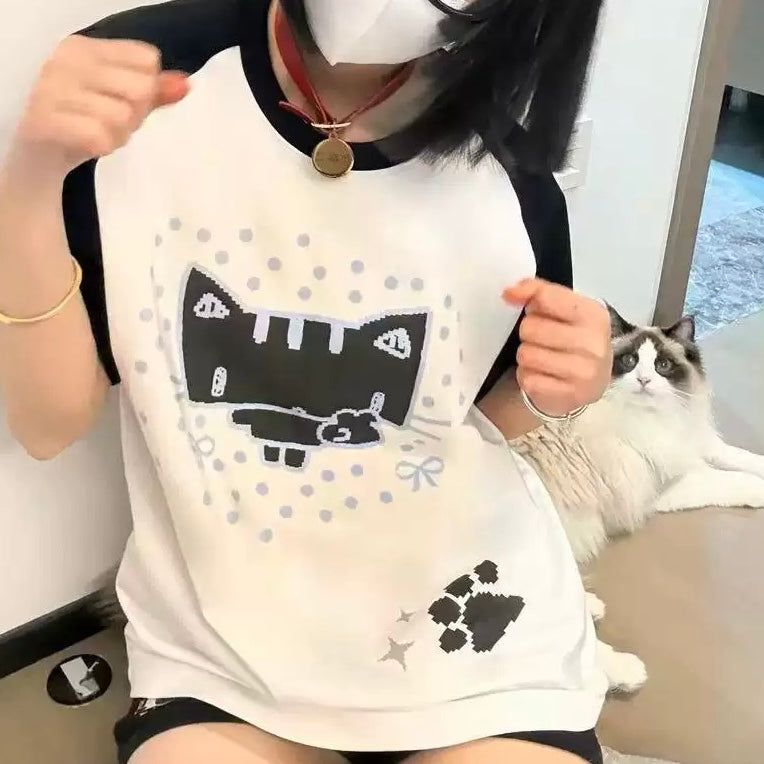 A person wearing a white and black raglan t-shirt with a black cat graphic, holding up the shirt to show the front. A fluffy white and gray cat is sitting on the floor in the background.