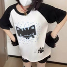 A person wearing a black and white raglan t-shirt with a pixelated cat graphic, black shorts, and black fuzzy wrist warmers, standing indoors.