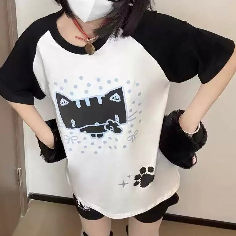 A person wearing a black and white raglan t-shirt with a pixelated cat graphic, black shorts, and black fuzzy wrist warmers, standing indoors.