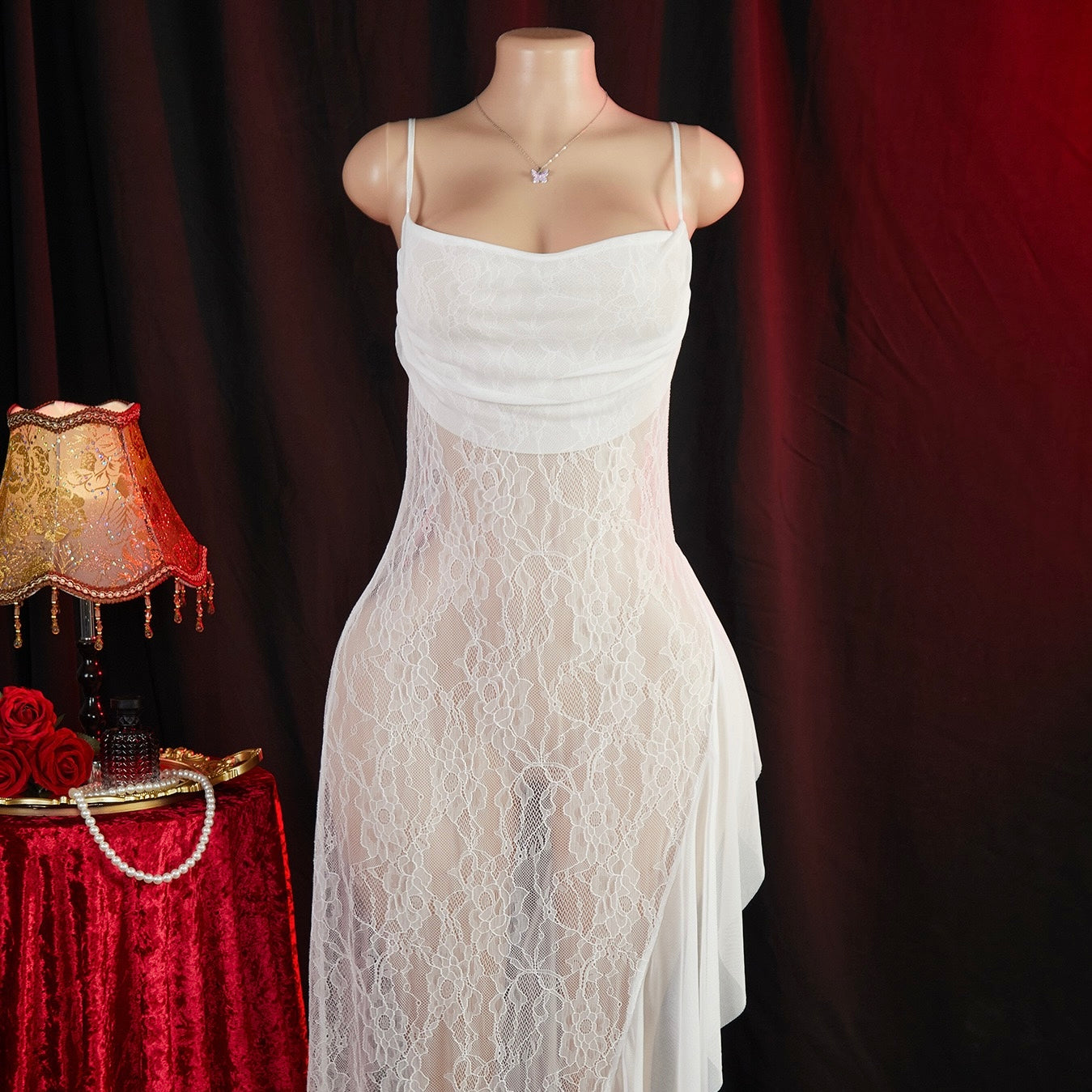 White lace dress on a mannequin against a dark background with a lamp and roses on a red table.
